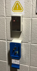 Metal panel at top of image with a small red and green light, with red light on it with the red light lit, and a blue lever panel is below metal panel with the pull lever pulled to the engaged position to lock doors.