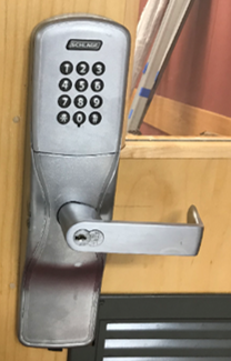 External view of metal lever door handle with with a numeric key pad  above the handle attached to a wooden door with a window at top of the door and a vent below